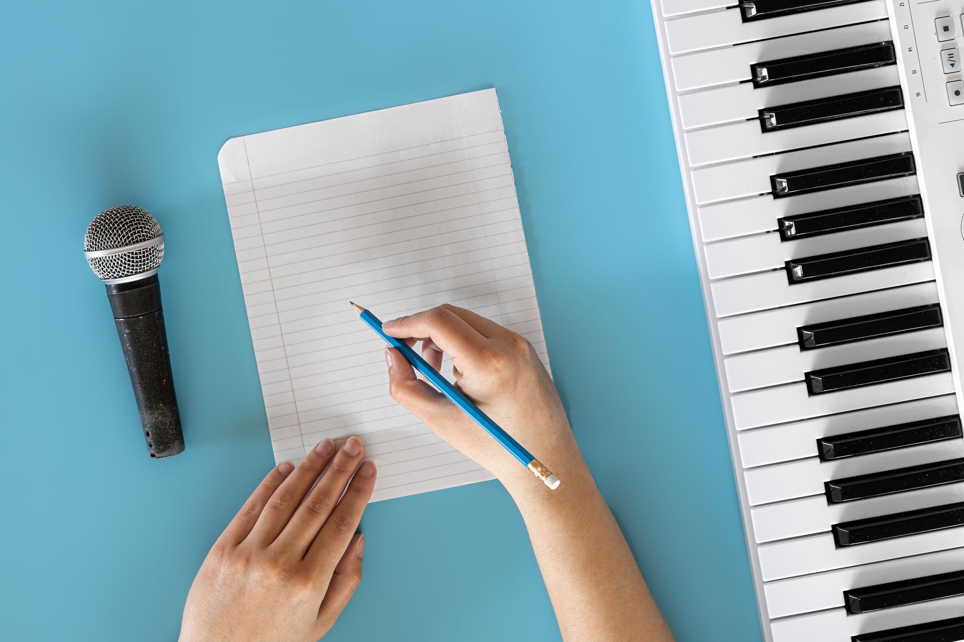 Person writing on notebook with a pencil next to microphone and keyboard on blue background.